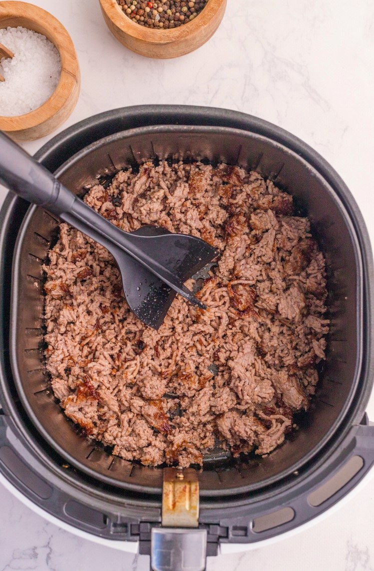 Cooked ground beef being stirred with a black spatula inside an air fryer basket, with bowls of salt and peppercorns visible in the background on a white countertop.