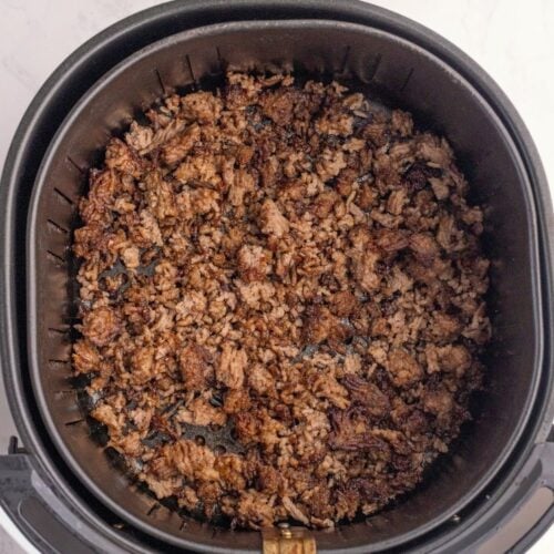Cooked and browned ground beef crumbles in an air fryer basket, viewed from above. Two small wooden bowls containing salt and pepper sit on a white marble surface near the air fryer.