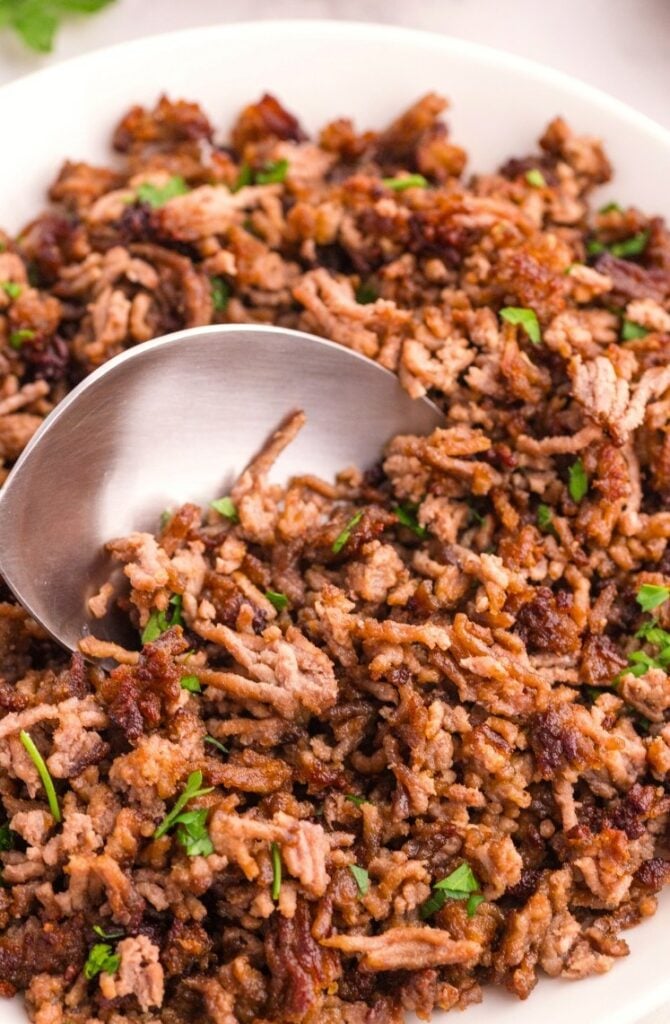 A close-up of cooked ground beef garnished with chopped parsley, served in a white dish with a metal serving spoon.