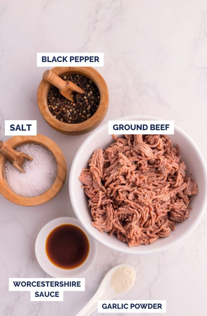 A bowl of ground beef on a marble surface, surrounded by small bowls of black pepper, salt, Worcestershire sauce, and garlic powder, each labeled with text.