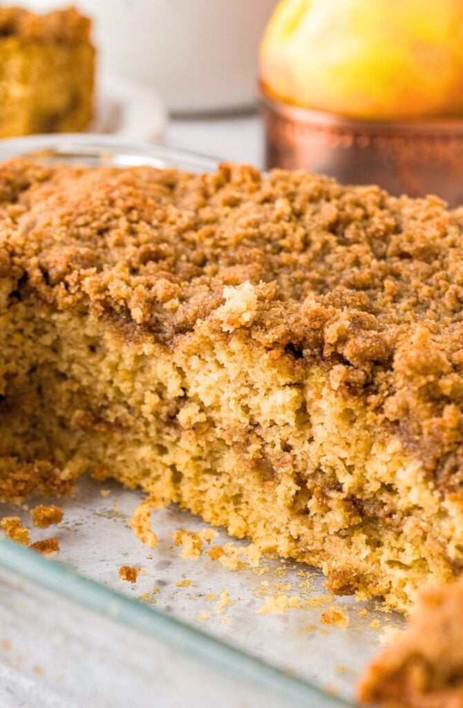 A close-up of a slice of coffee cake with a crumbly cinnamon streusel topping in a glass baking dish. A partial piece is missing, revealing the moist, fluffy interior. An apple is blurred in the background.