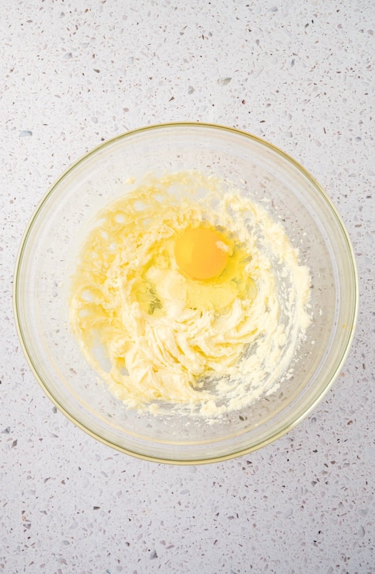 A glass bowl on a speckled countertop contains creamed butter and sugar with a raw egg cracked on top, ready to be mixed together.