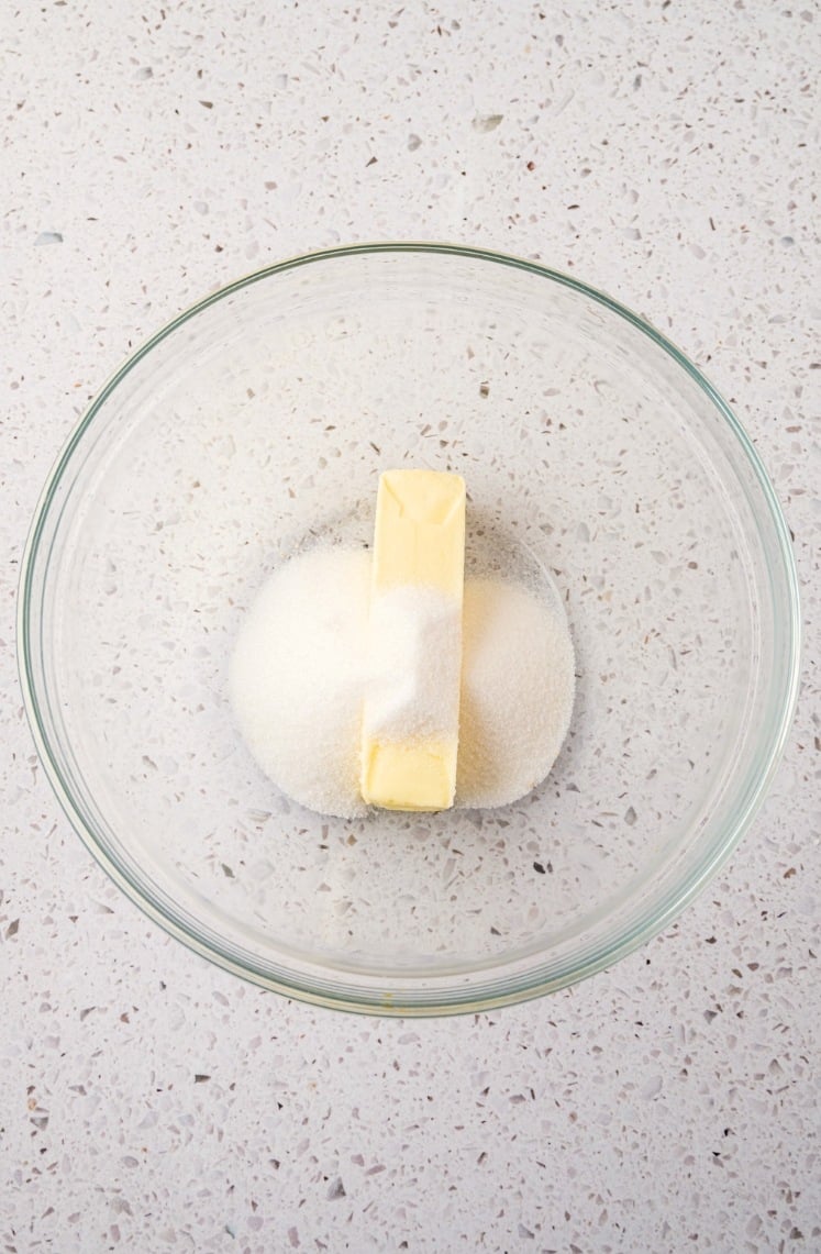 A glass bowl with a stick of butter and a pile of granulated sugar inside it, placed on a light-colored speckled countertop.