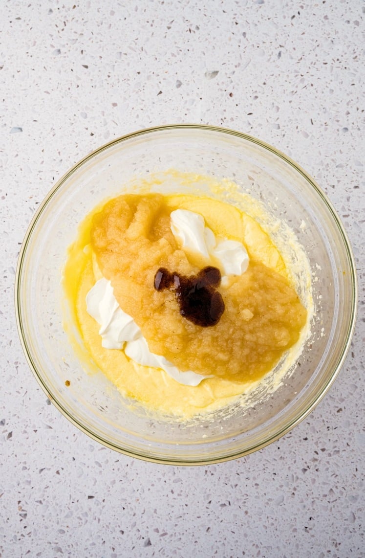 A glass mixing bowl containing a yellow batter, with dollops of white cream, a portion of applesauce, and a splash of vanilla extract on top, all sitting on a speckled countertop.