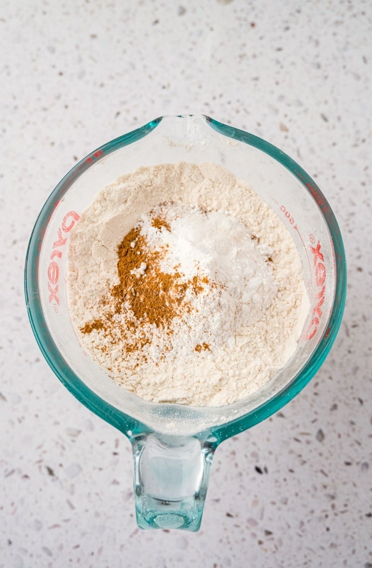 A glass measuring cup contains flour, baking powder, and ground spices on a speckled countertop, ready to be mixed for baking.