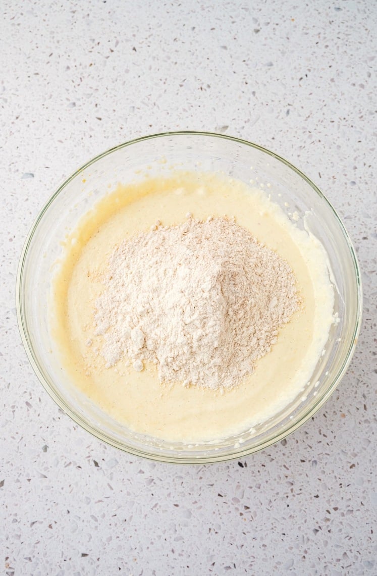 A glass bowl containing cake batter with a mound of dry flour on top, sitting on a speckled countertop.