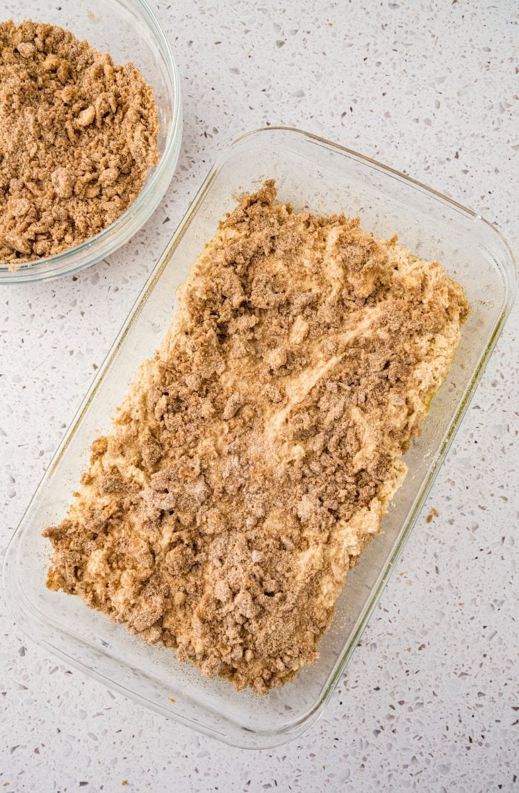 A glass baking dish filled with an unbaked crumb cake topped with a brown sugar and cinnamon crumble, sits on a light speckled countertop. A bowl of extra crumble topping is nearby.