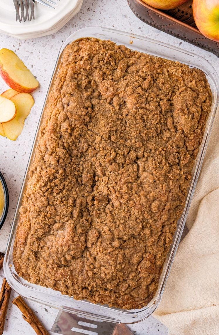 A glass baking dish filled with a golden-brown crumb-topped dessert, possibly apple crisp or coffee cake, sits on a countertop near sliced apples, cinnamon sticks, and a beige napkin.