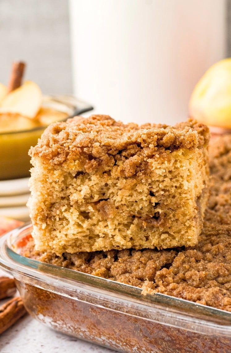 A thick slice of crumb-topped coffee cake sits on top of the rest of the cake in a glass baking dish, showing a moist, golden interior with a cinnamon crumb topping.