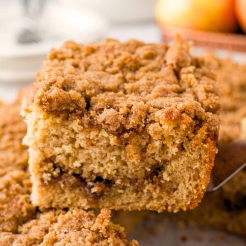 A close-up of a slice of cinnamon coffee cake with a crumbly streusel topping being lifted from a tray, with apples and a glass of milk blurred in the background.