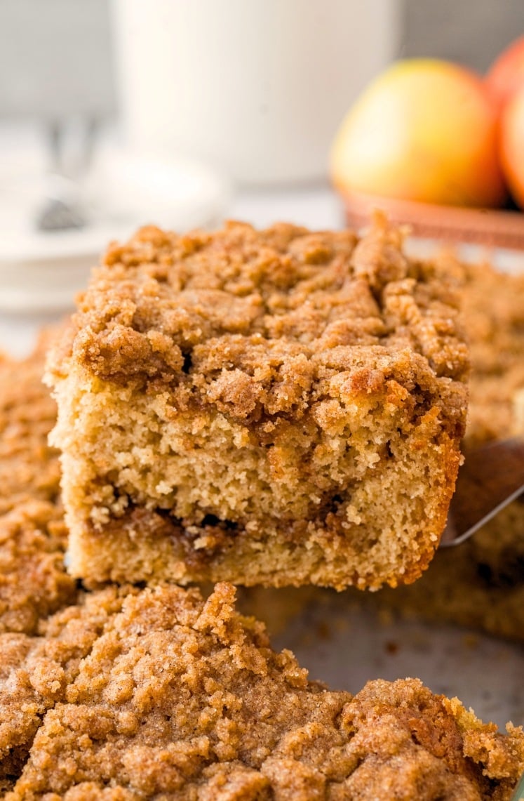 A close-up of a slice of cinnamon coffee cake with a crumbly streusel topping being lifted from a tray, with apples and a glass of milk blurred in the background.