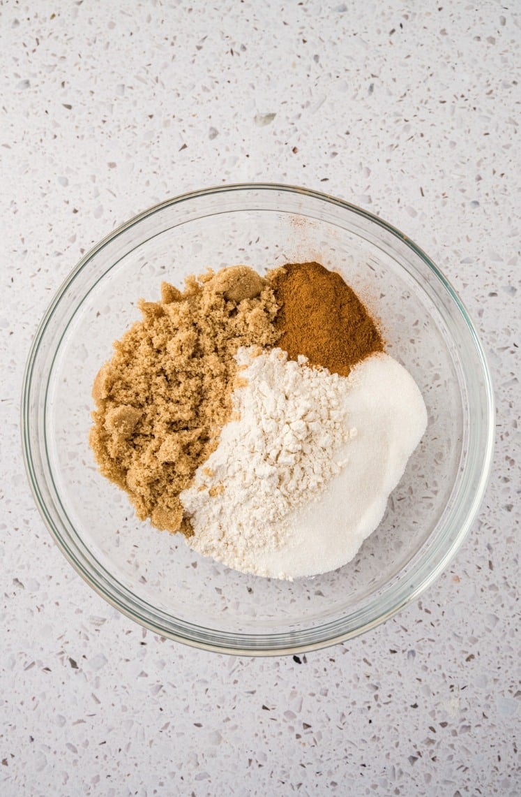 A glass bowl containing brown sugar, white sugar, flour, and cinnamon, arranged in separate piles on a speckled countertop.
