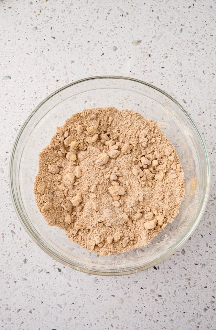 A clear glass bowl containing a crumbly mixture of brown sugar and flour sits on a light-colored speckled countertop.