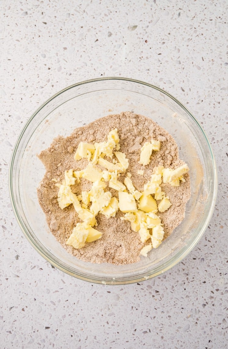 A glass bowl on a speckled countertop containing a mixture of brown sugar and flour, topped with chunks of butter before mixing.