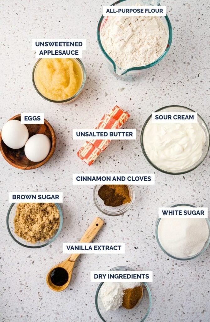 Overhead view of labeled baking ingredients on a countertop: all-purpose flour, unsweetened applesauce, eggs, unsalted butter, sour cream, brown sugar, cinnamon and cloves, white sugar, vanilla extract, and dry ingredients.