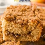 A close-up of a slice of crumbly applesauce coffee cake being lifted from a tray, with a cinnamon streusel topping and a soft, moist interior. Text on the image reads "Applesauce Coffee Cake real balanced.