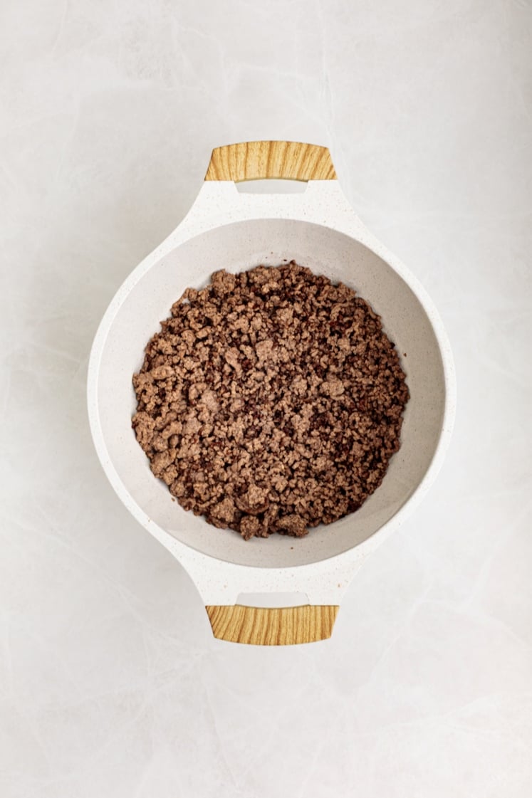 A white pot with wooden handles contains browned ground beef, ready for making Baked Beans with Ground Beef, viewed from above on a light-colored surface.