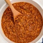A white pot filled with Baked Beans with Ground Beef and savory sauce is being stirred with a wooden spoon. A striped kitchen towel and a salt shaker sit nearby on the counter.