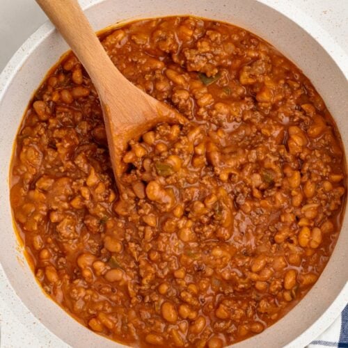 A white pot filled with Baked Beans with Ground Beef and savory sauce is being stirred with a wooden spoon. A striped kitchen towel and a salt shaker sit nearby on the counter.