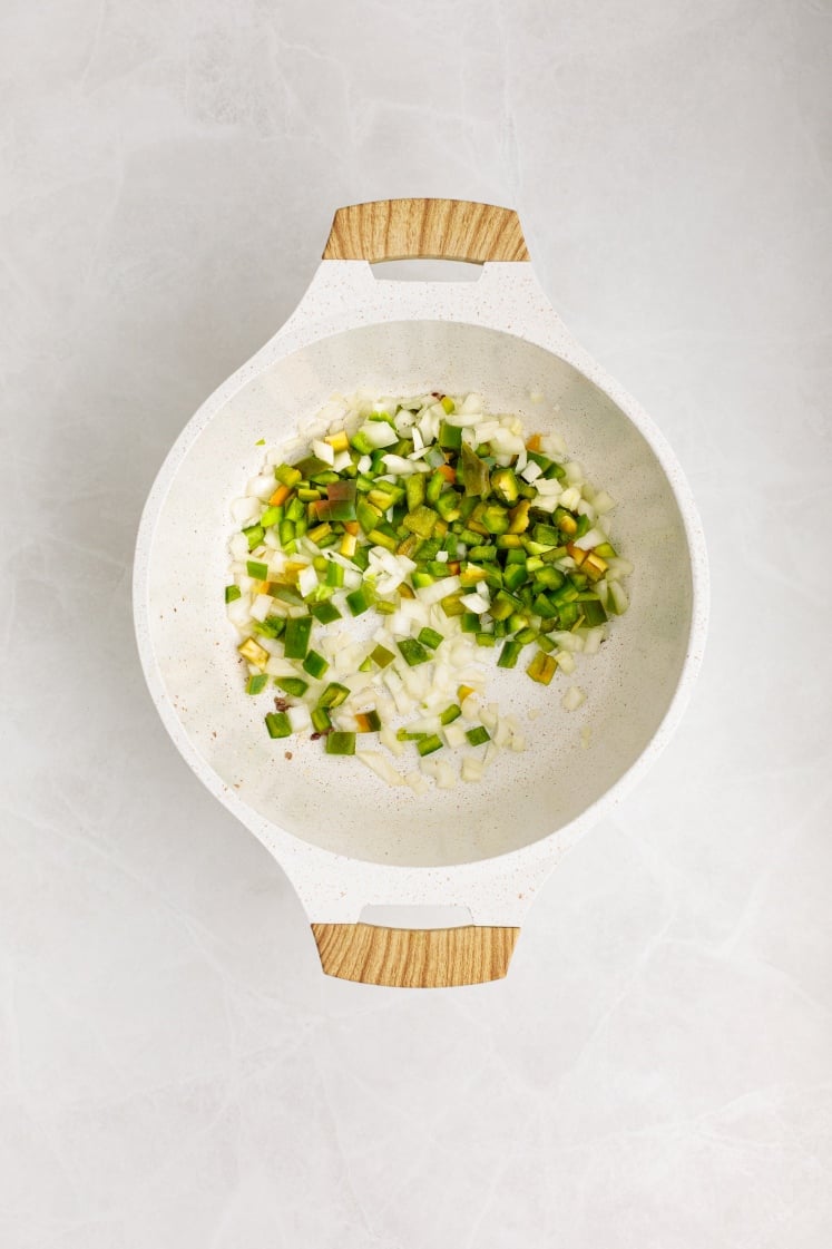 A white pot with wooden handles contains chopped green bell peppers, onions, and garlic pieces—perfect starter ingredients for hearty Baked Beans with Ground Beef—resting on a light countertop.