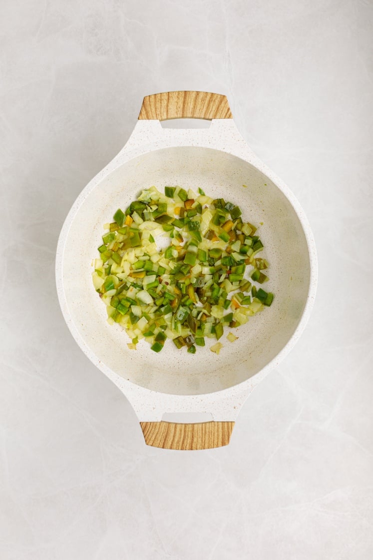 A white pot with wooden handles contains chopped green and yellow vegetables, possibly zucchini and peppers, perfect as a fresh side for Baked Beans with Ground Beef, on a light marble surface.
