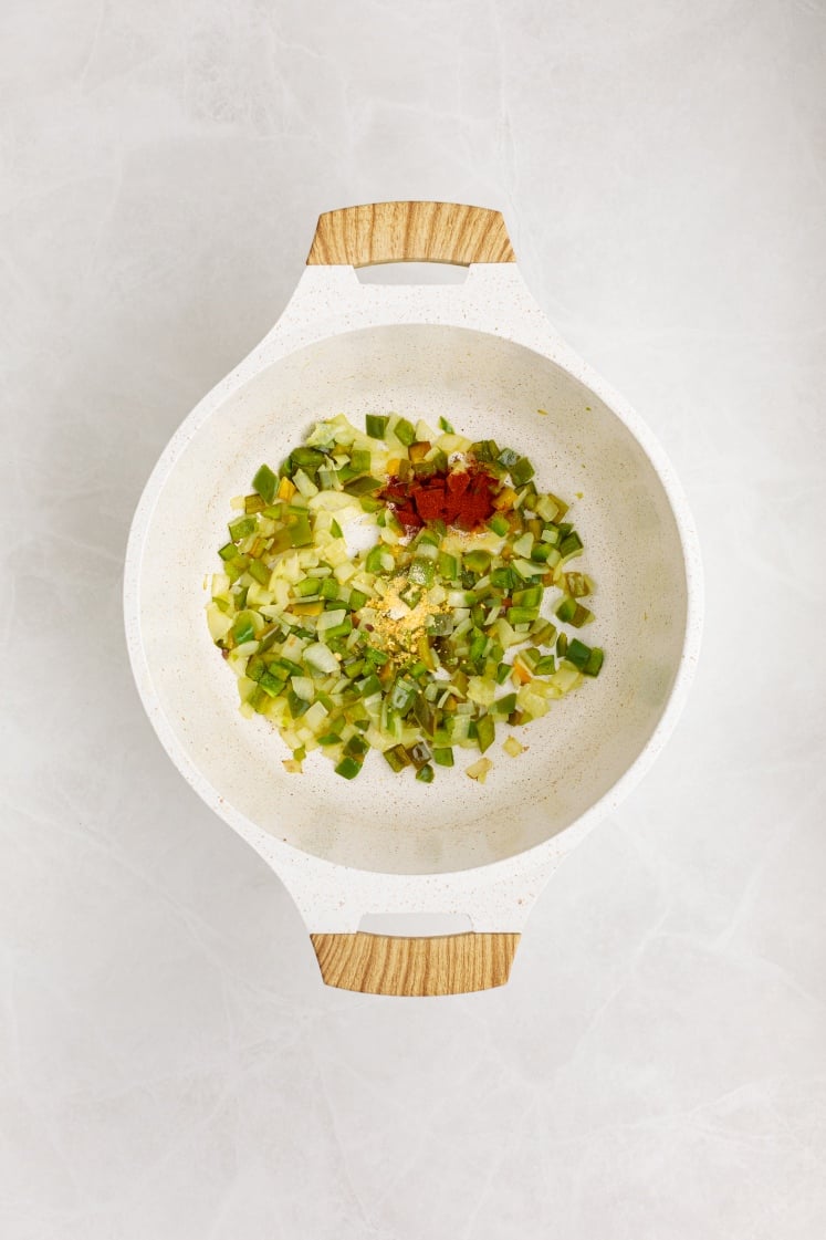 A white pot with wooden handles contains chopped green vegetables, minced garlic, and a sprinkle of red and yellow spices—perfect starters for making hearty Baked Beans with Ground Beef—resting on a light-colored marble surface.