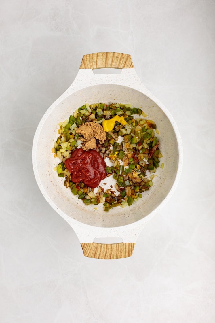 A white pot with light wooden handles holds chopped sautéed vegetables, Baked Beans with Ground Beef, ketchup, mustard, and brown sugar, all sitting atop a light-colored surface.