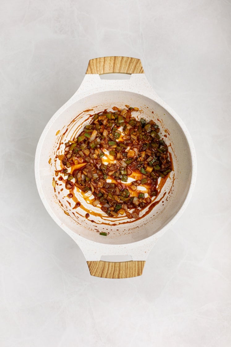 A white pot with wooden handles contains sautéed vegetables, spices, and Baked Beans with Ground Beef in a rich reddish sauce, sitting on a light-colored countertop.