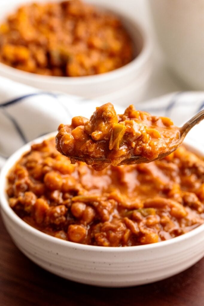 A close-up of a white bowl filled with baked beans in a thick, rich sauce. A spoonful of the beans is being held above the bowl, showing their hearty texture. Another bowl of beans is blurred in the background.