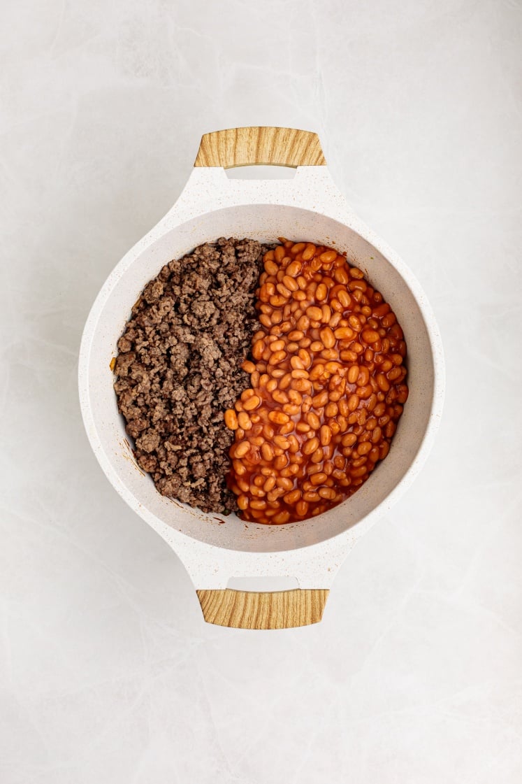 A round white pot with wooden handles contains Baked Beans with Ground Beef—cooked ground beef on one side and savory baked beans in sauce on the other—placed on a light-colored surface.