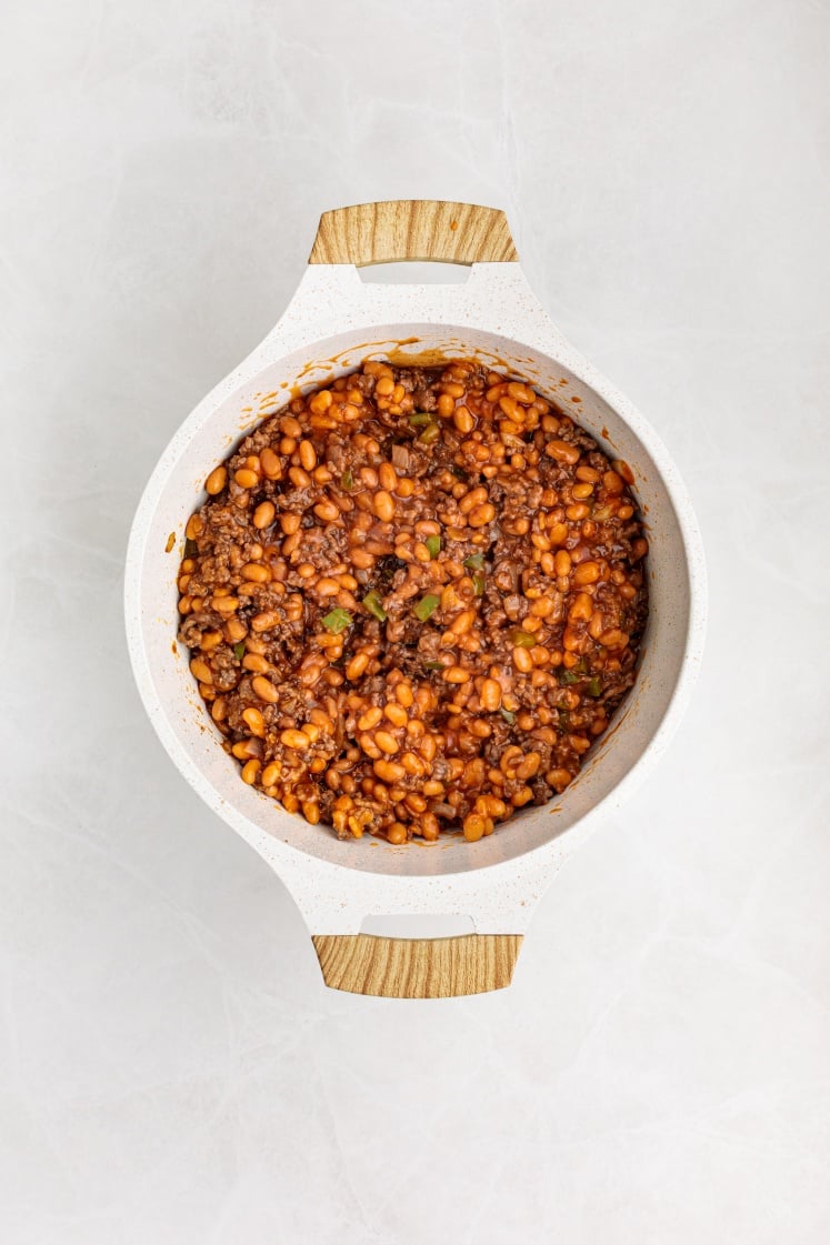 A white pot with light wooden handles filled with Baked Beans with Ground Beef in a rich tomato sauce, viewed from above on a light background.