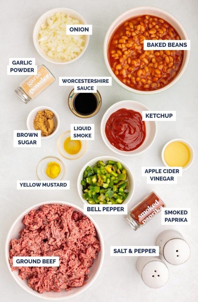 Overhead view of labeled ingredients for Baked Beans with Ground Beef: baked beans, ketchup, Worcestershire sauce, liquid smoke, apple cider vinegar, yellow mustard, brown sugar, garlic powder, onion, bell pepper, smoked paprika, salt, and pepper.