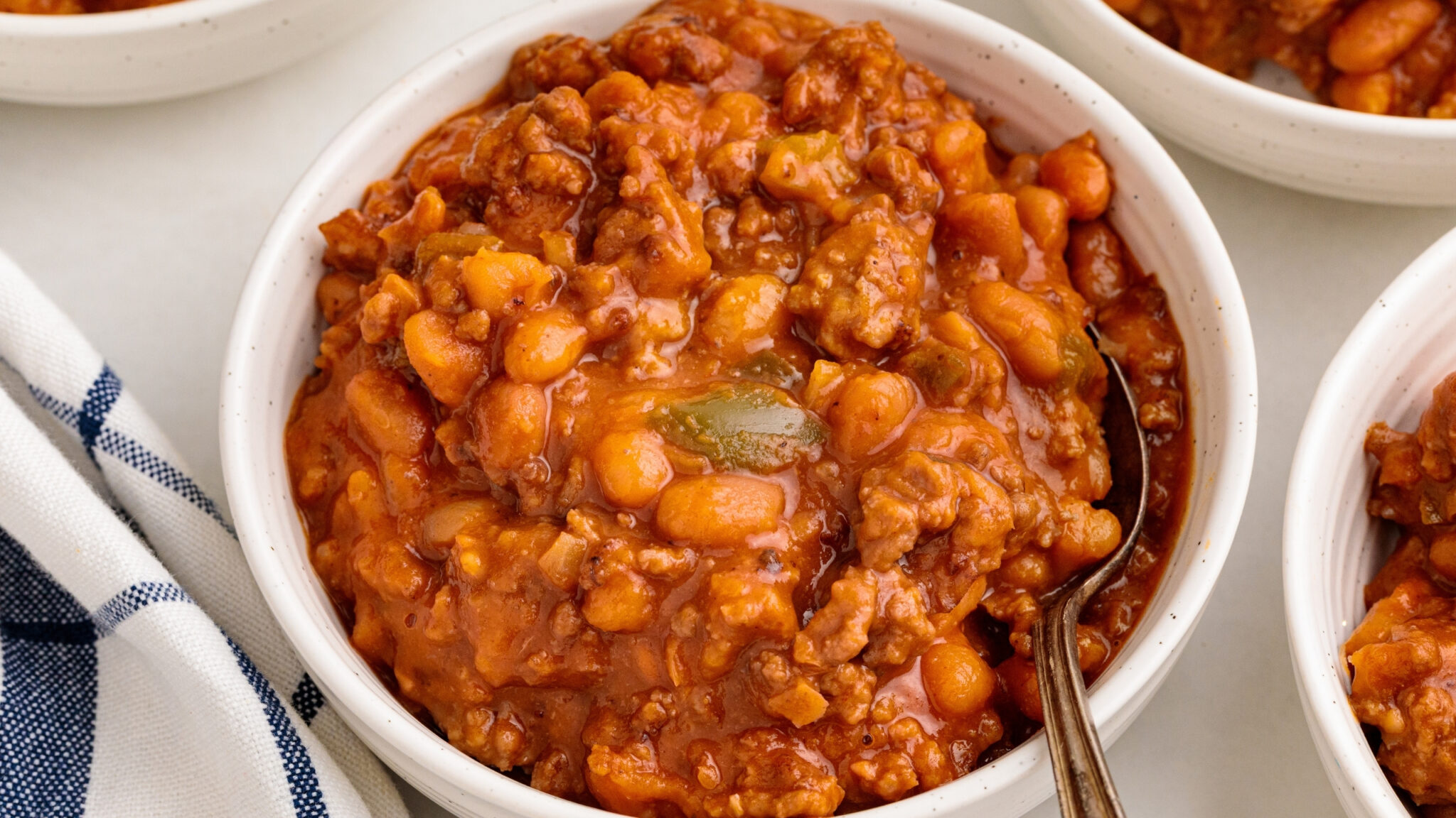 A pot of ground beef and beans being stirred with a wooden spoon, set on a wooden surface next to a blue and white striped cloth and a salt shaker.