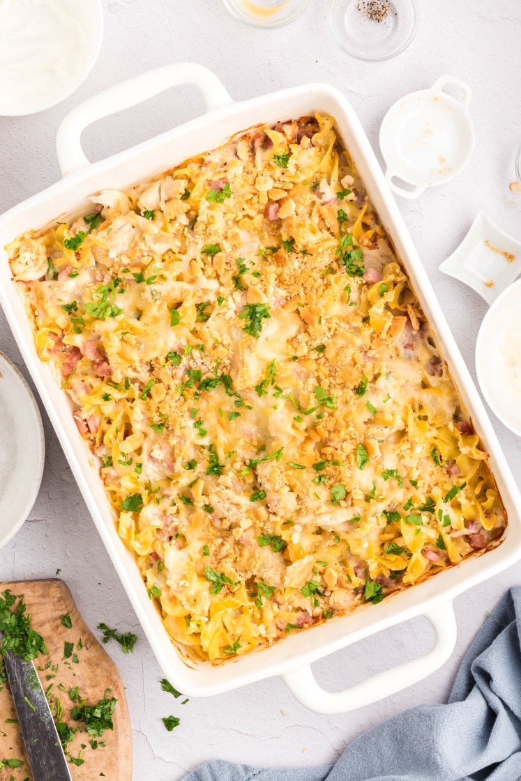 A baked casserole in a white rectangular dish, topped with melted cheese, bread crumbs, and chopped parsley. Pieces of pasta and bits of ham or chicken are visible. The dish is surrounded by bowls and utensils on a light surface.