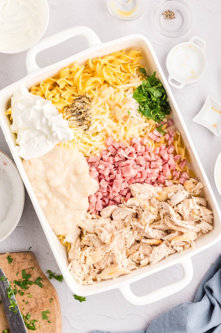 A white baking dish filled with uncooked egg noodles, shredded chicken, diced ham, shredded cheese, chopped parsley, sour cream, condensed soup, and seasonings, all arranged in separate piles.