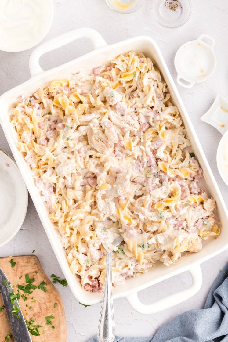 A white baking dish filled with creamy chicken tetrazzini pasta, featuring noodles, chicken pieces, and a creamy sauce, sitting on a light-colored surface with a spoon in the dish and bowls nearby.
