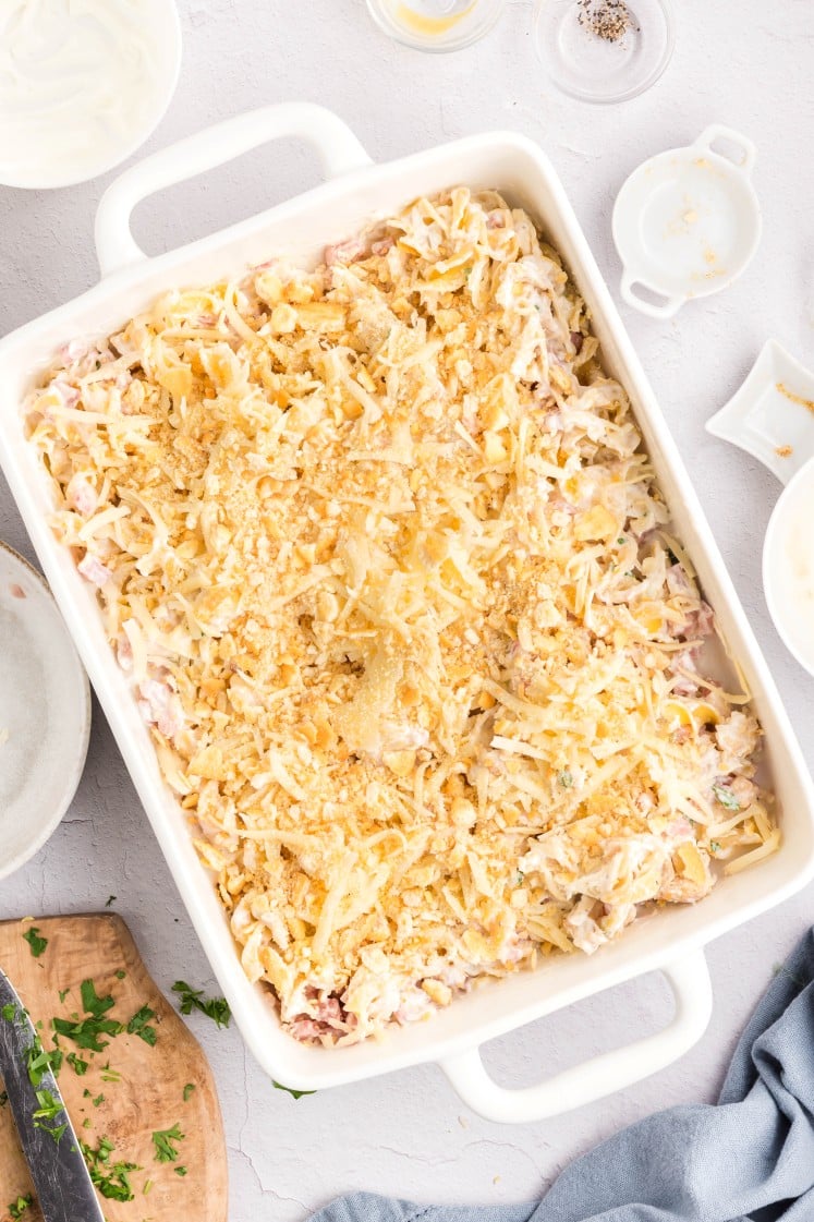 A white baking dish filled with an unbaked casserole topped with shredded cheese and crushed crackers, surrounded by empty dishes and food prep items on a light-colored countertop.