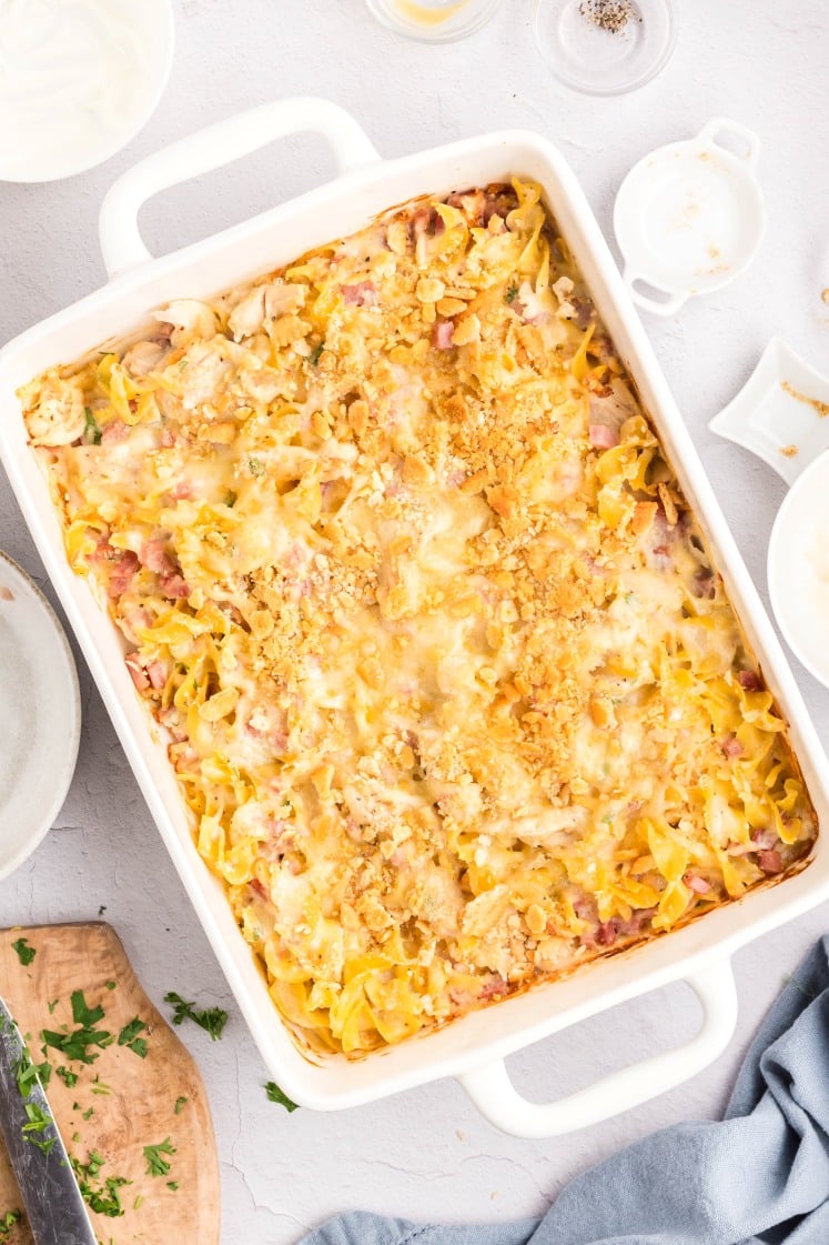 A baked casserole with noodles, cheese, and a golden breadcrumb topping sits in a white rectangular dish on a light surface, surrounded by small bowls and a folded blue cloth.