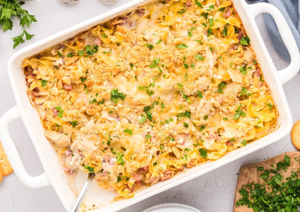 A baked casserole in a white dish, topped with melted cheese, breadcrumbs, and chopped parsley. A serving spoon rests inside, and some noodles and ingredients are visible around the edges.