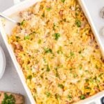 A white baking dish filled with a cheesy noodle casserole topped with chopped parsley and crushed crackers, with a serving spoon resting in the dish. Crackers and fresh herbs are visible nearby.
