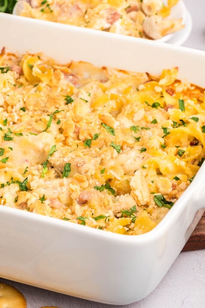 A close-up of a baked casserole in a white dish, topped with golden breadcrumbs and chopped herbs, featuring visible pieces of chicken and pasta.
