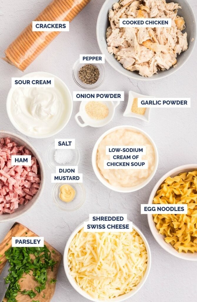 Ingredients for a casserole are arranged on a white surface, including bowls of cooked chicken, egg noodles, ham, shredded Swiss cheese, sour cream, cream of chicken soup, spices, parsley, dijon mustard, and a stack of crackers.