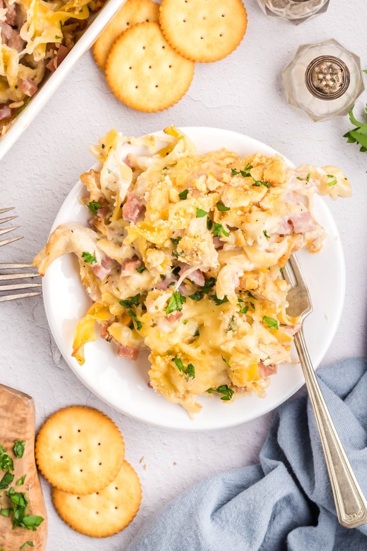 A plate of Chicken Cordon Bleu Casserole with cheesy noodles, bits of ham, and parsley sits on a white table surrounded by round crackers, a blue napkin, and a fork.