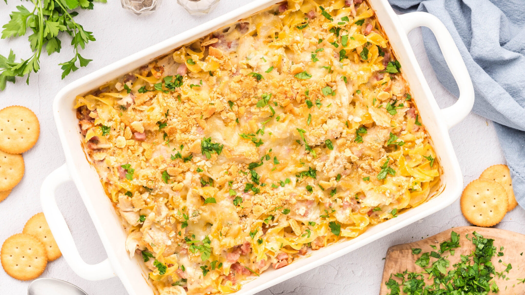 A white baking dish filled with chicken cordon bleu casserole, topped with melted cheese, crushed crackers, and chopped parsley.