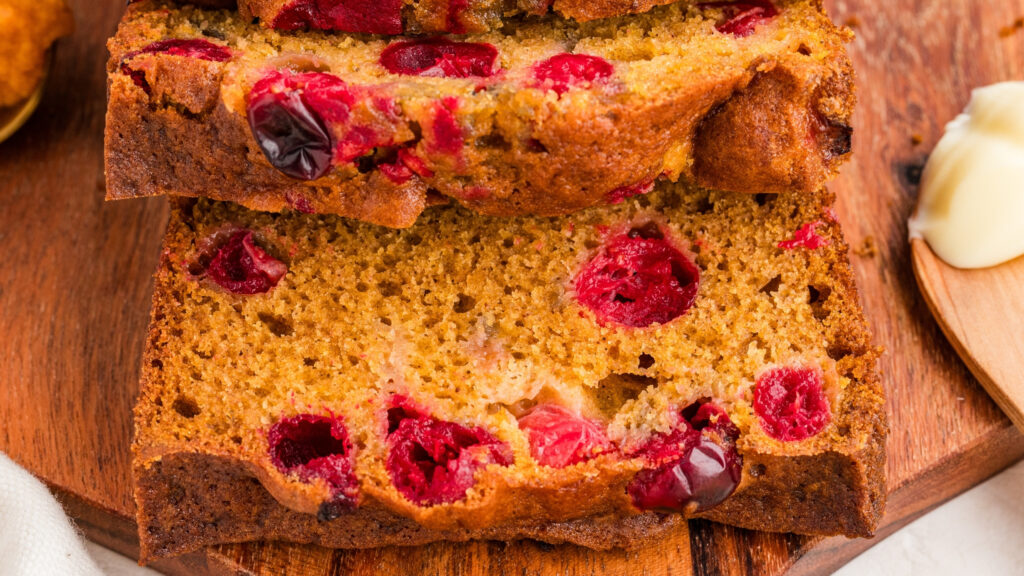 Two thick slices of pumpkin cranberry bread rest on a wooden board, showing the tender, moist interior filled with whole cranberries and a golden pumpkin-hued crumb.