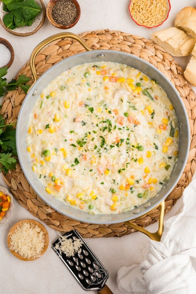 A large skillet filled with creamy orzo pasta topped with chopped parsley. Ingredients like corn, peas, carrots, grated cheese, and seasonings are arranged around the skillet on a woven placemat.