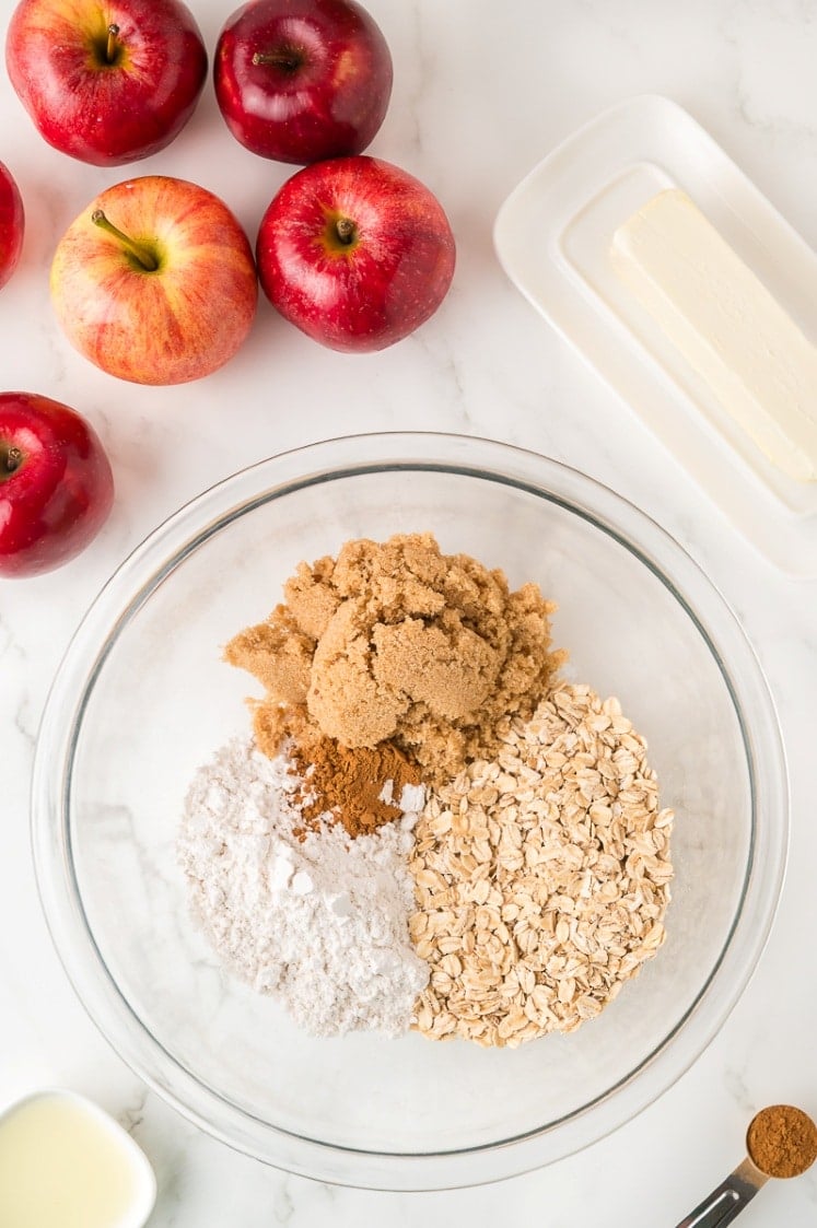 A glass bowl contains oats, brown sugar, flour, and cinnamon. Surrounding the bowl are several red apples, a stick of butter on a dish, and a small bowl of liquid, all on a white surface.
