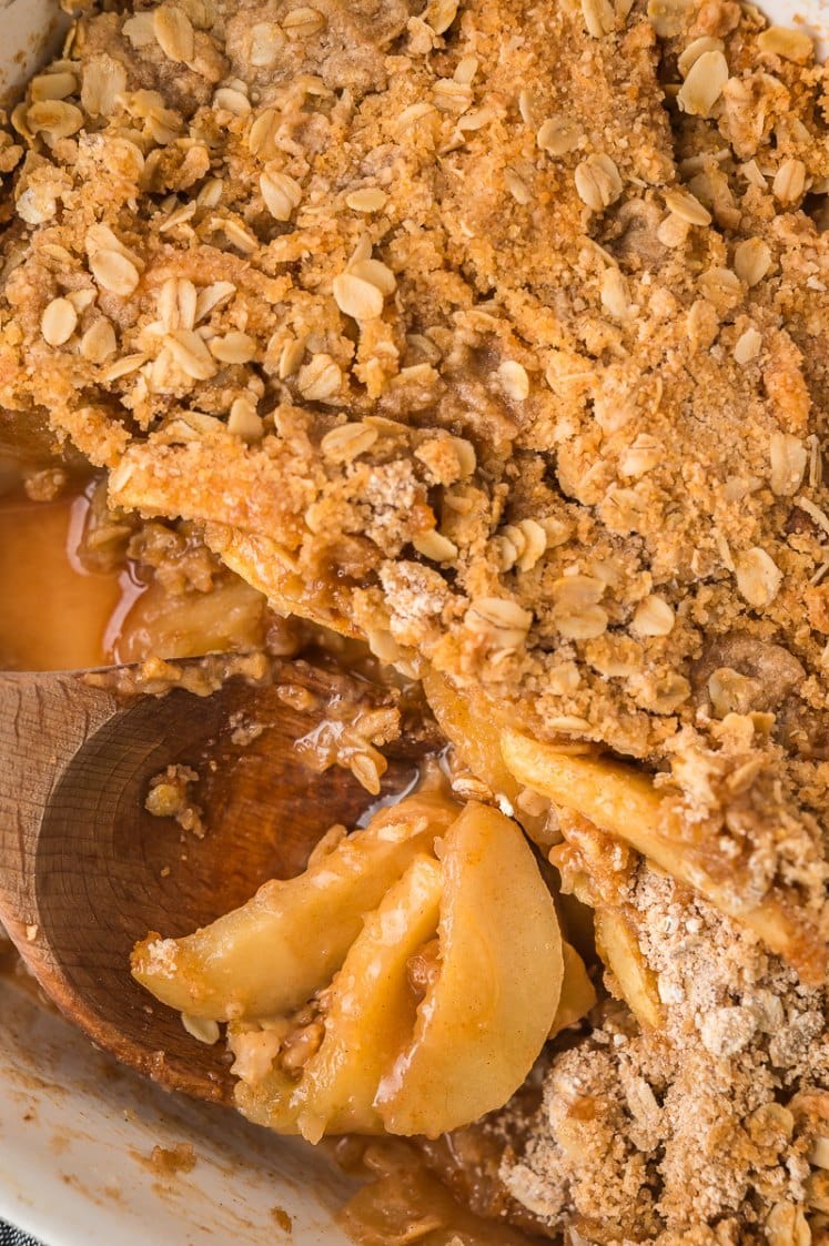 Close-up of a baked apple crisp with golden oat and crumb topping, revealing tender, cinnamon-spiced apple slices underneath. A wooden spoon is scooping a portion from the baking dish.