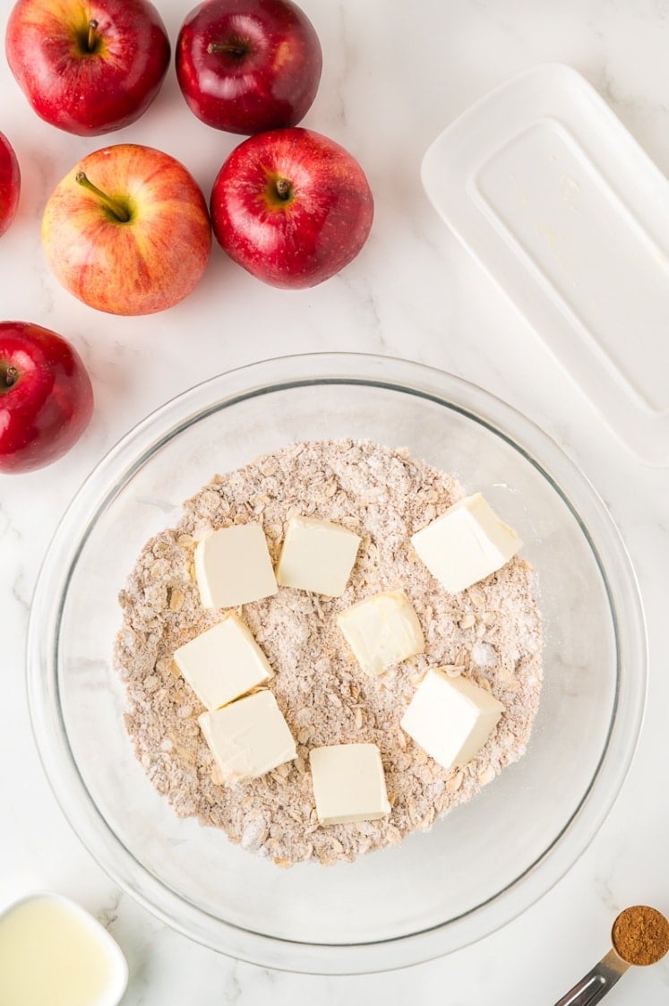 A glass bowl with flour mixture and cubes of butter sits on a white surface, surrounded by several red apples, a small pitcher of liquid, a white dish, and a small bowl of ground spice.