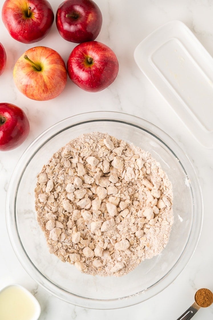 A glass bowl filled with crumbly flour mixture sits on a white surface, surrounded by whole red apples, a small dish of melted butter, a spice, and a white baking dish.